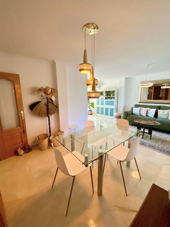 A modern living room with a glass dining table, white chairs, a green sofa, and a ceiling light fixture.