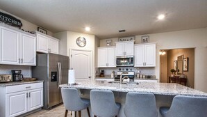 A modern kitchen with white cabinetry, a stainless steel refrigerator, a built-in oven, and a large island with bar stools.