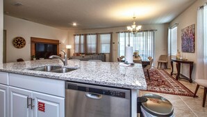 A modern kitchen with granite countertops, stainless steel appliances, and a dishwasher. There is a dining area with a chandelier, a rug, and a painting on the wall.