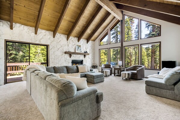 Cozy living room with a high ceiling, fireplace, and large windows offering views of the surrounding forest.
