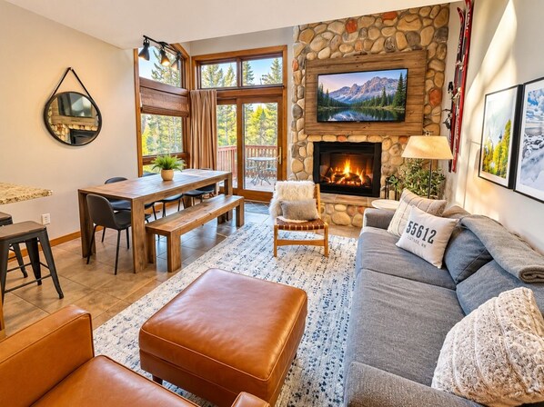 A cozy living room with a stone fireplace, a television mounted above it, and a large window offering a view of the outdoors.