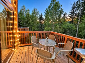 Une terrasse en bois avec une table et des chaises en verre, entourée d’une rambarde, donnant sur une zone boisée.