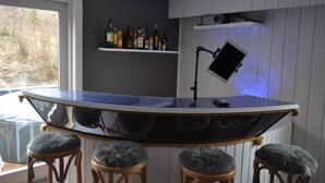 A bar area with a curved counter, a mounted TV, and a shelf with bottles. There are three stools with fur-cushioned seats.