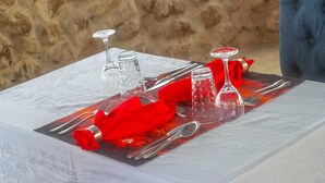 Une table dressée avec de la vaisselle en verre transparent, une serviette rouge et des couverts en argent, le tout sur fond d’un mur en pierre.