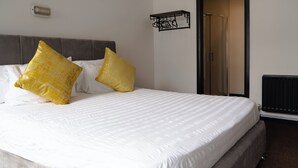 A neatly made bed with white bedding and two large yellow pillows, a gray upholstered headboard, a black-framed mirror, and a black radiator. - Bushmills Hostel (Bushmills)