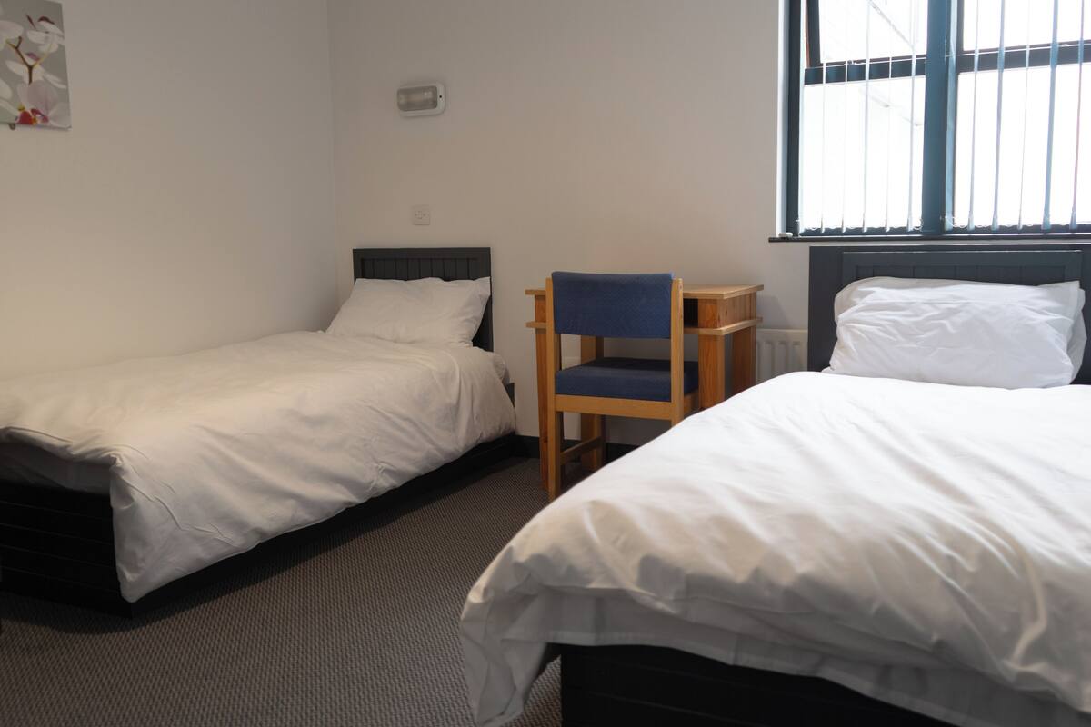 A room with two single beds, a wooden chair, and a window with blinds.