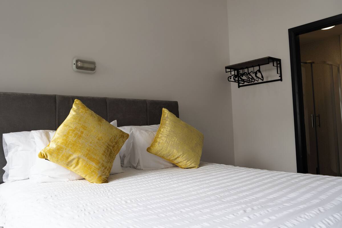 A neatly made bed with white bedding and two large gold pillows, a gray headboard, and a small black shelf with a geometric design on the wall.