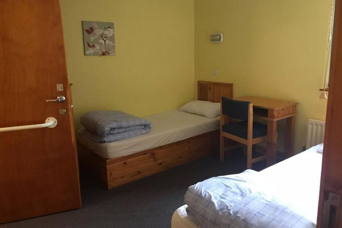 A single bed with a wooden frame, a small desk, and a chair in a room with yellow walls.