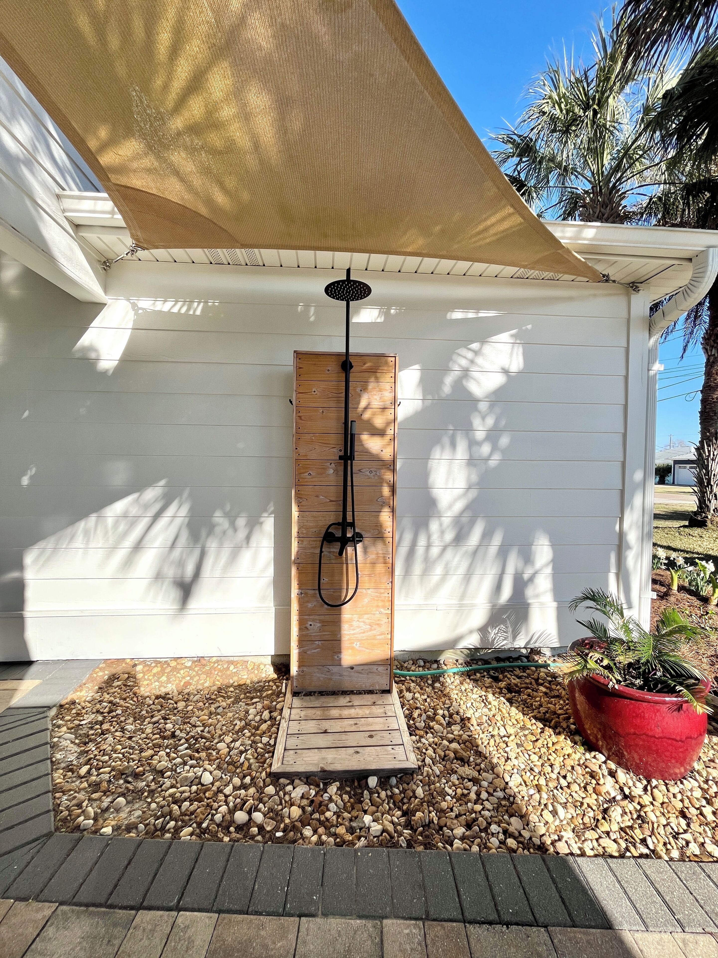 A wooden outdoor shower with a hose, surrounded by pebbles and a red planter.