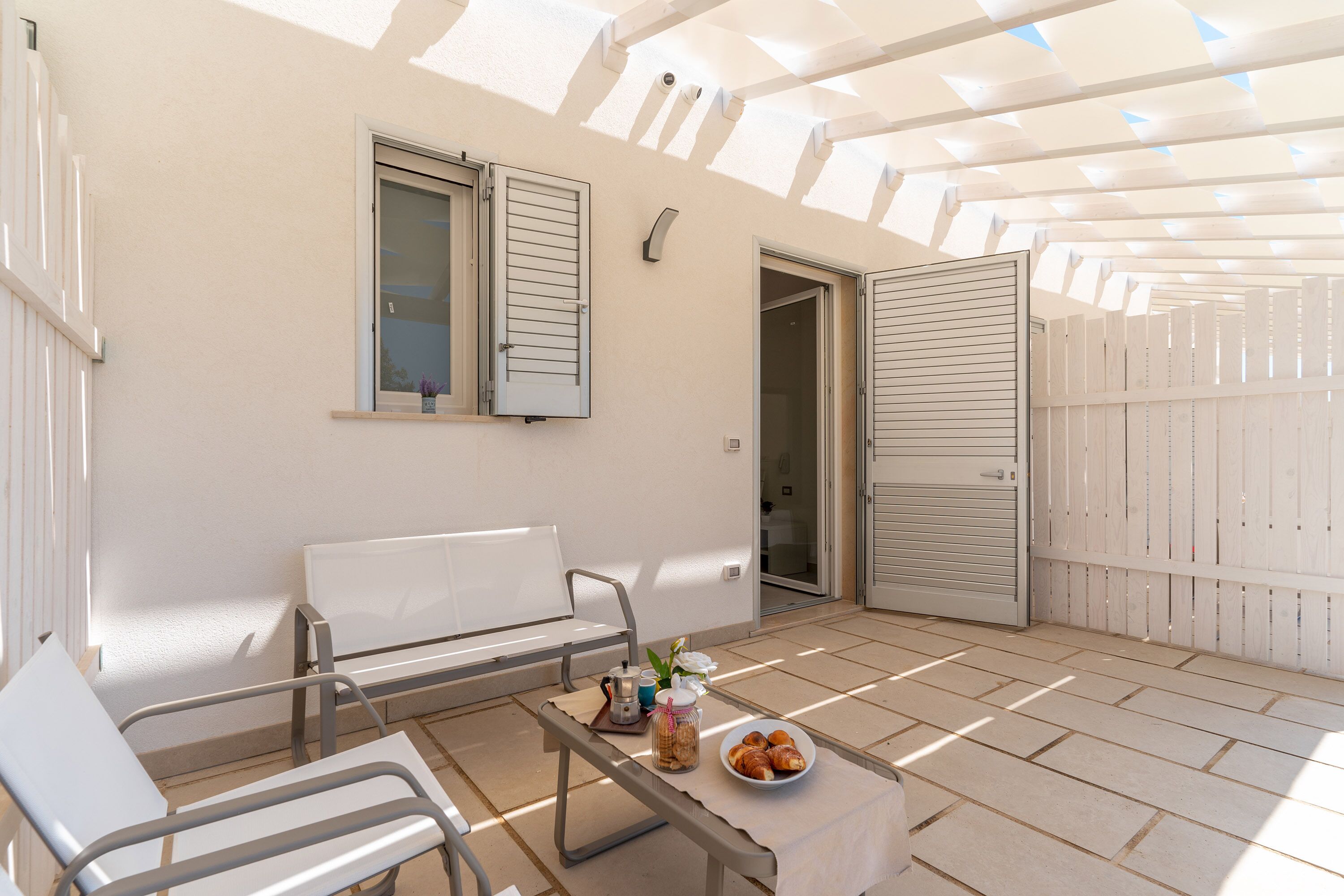 Un patio soleado con un banco blanco, una mesa con un bol de pasteles y una puerta que da a un espacio interior.