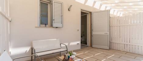 Un patio soleado con un banco blanco, una mesa con un bol de pasteles y una puerta que da a un espacio interior.