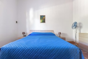 Ein Zimmer mit einem Bett, einem Wandbild, einem Standventilator und Nachttischlampen.