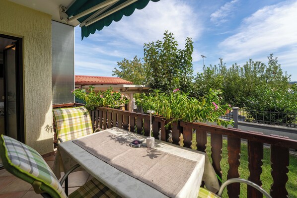 A balcony with a table and chairs, a pergola, and a view of greenery and a building in the background.