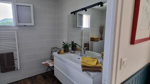Un baño con pared de azulejos blancos, una ventana, toallero, lavabo con espejo y una pequeña planta en maceta.
