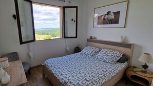 Un dormitorio con cama, ventana, mesita de noche, lámpara y un cuadro en la pared.