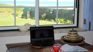 Un ordinateur portable est posé sur une table près d’une fenêtre, d’où l’on a vue sur un jardin et la mer.