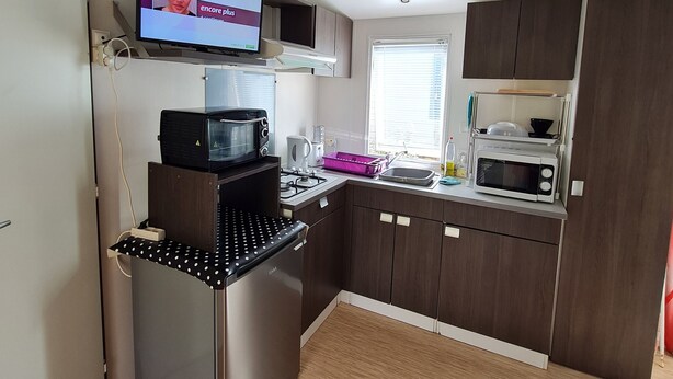 A compact kitchen with a built-in microwave, a small island with a kettle, and a flat-screen TV mounted above the island. - Mobile home rental (Mimizan)