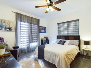 A bedroom with a bed, a ceiling fan, a window with blinds, a nightstand, and a lamp.
