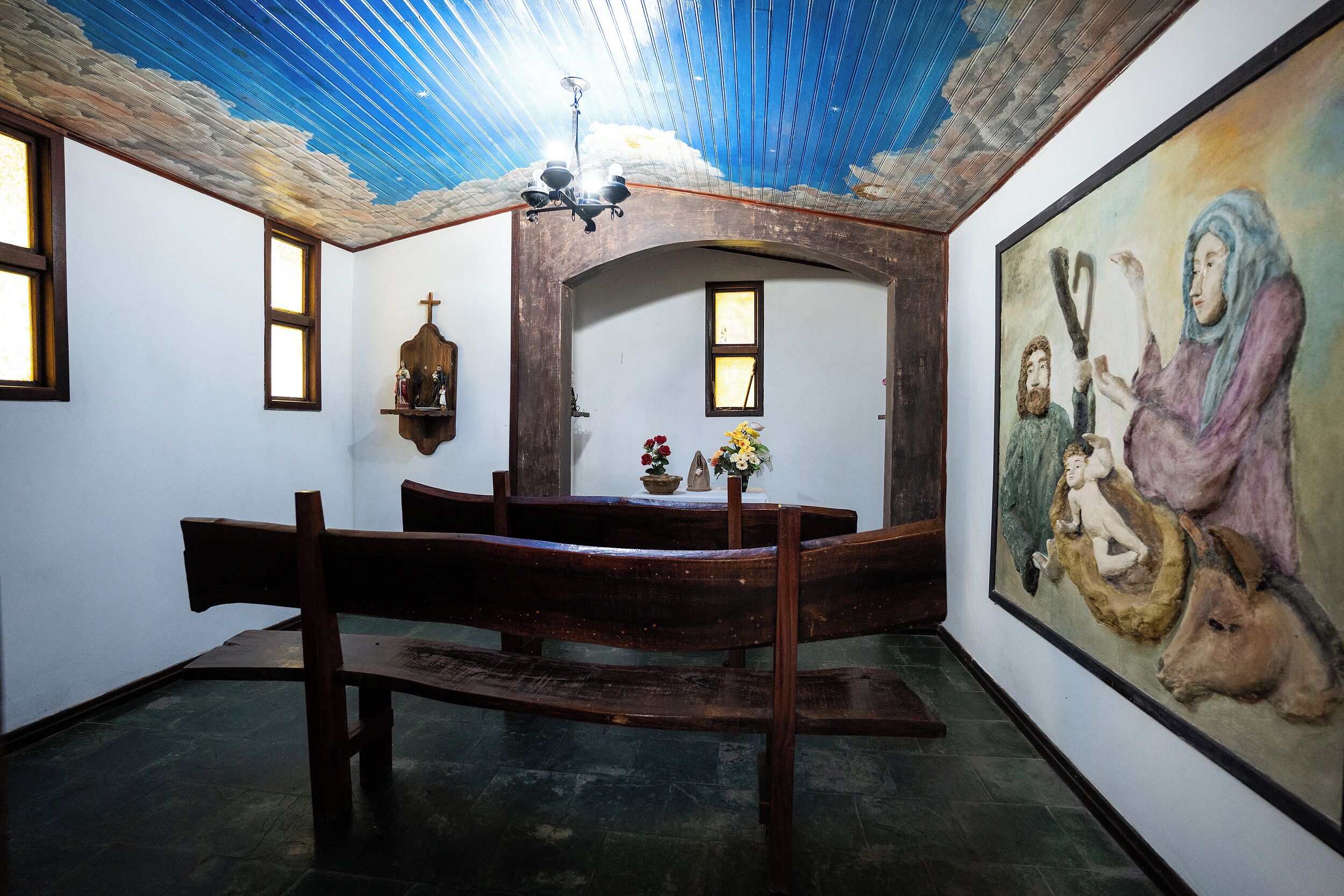 A church interior with wooden benches, a cross, and a painting on the wall.