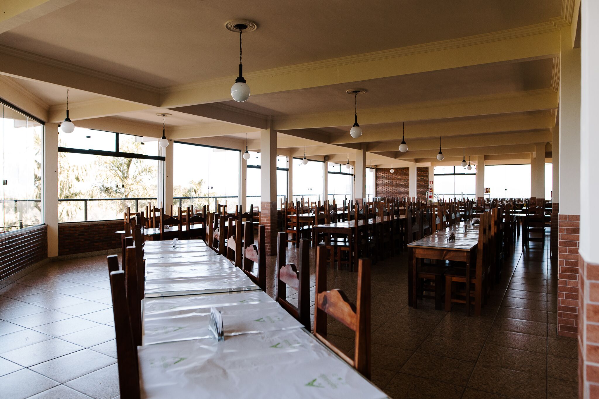 A large dining hall with long tables and chairs, large windows, and hanging pendant lights.