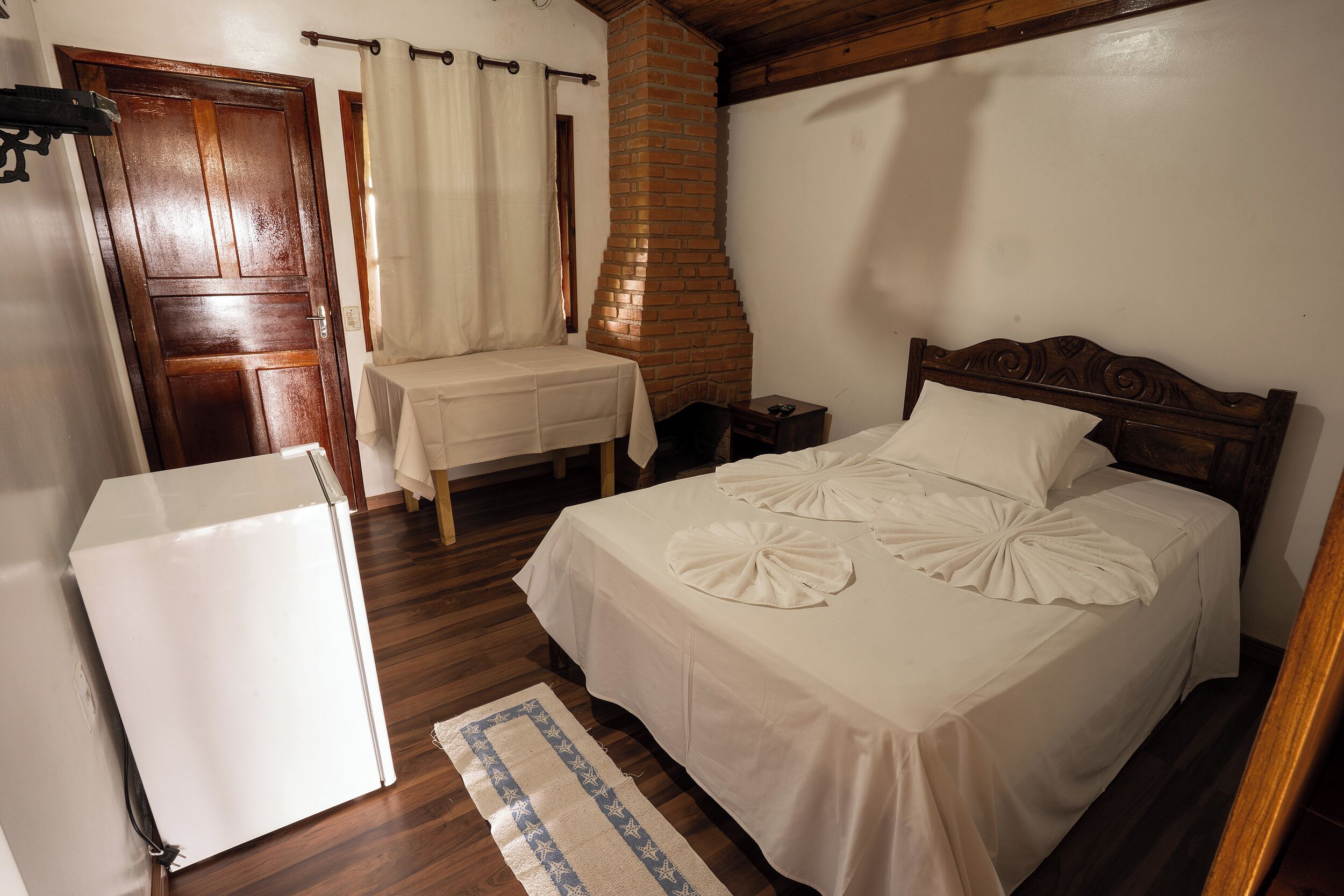 Une chambre à coucher comprenant un lit, une petite table, une chaise, une armoire en bois, un rideau blanc et une cheminée en briques.