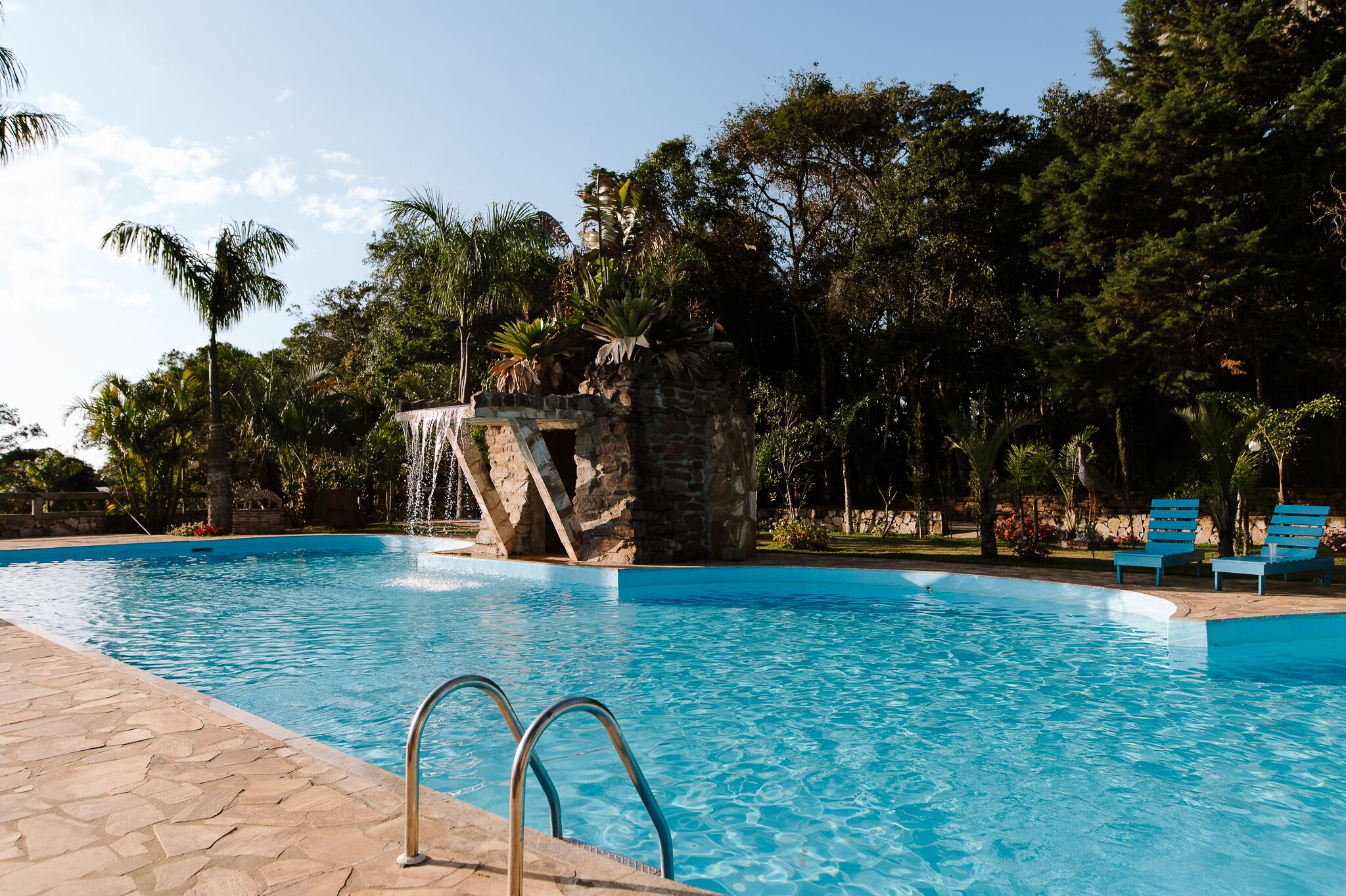 A swimming pool with a waterfall feature, surrounded by lush greenery and stone structures.