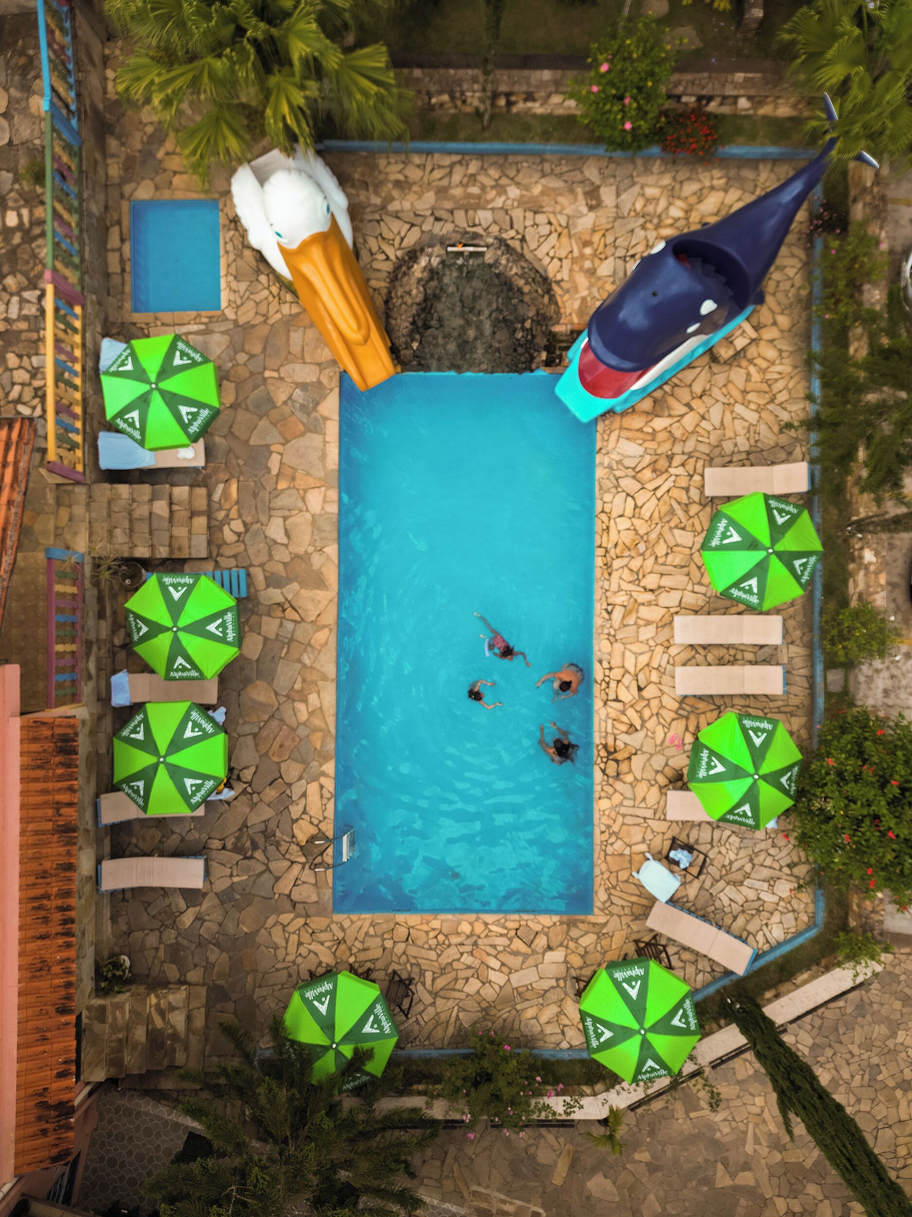 An aerial view of a swimming pool with a stone wall, surrounded by green umbrellas, and a water slide.