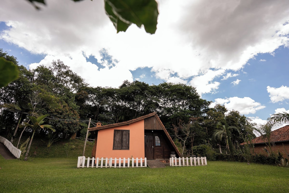 Uma casa pequena com telhado inclinado, cercada por uma cerca branca de piquete, situada em paisagem verdejante com árvores.