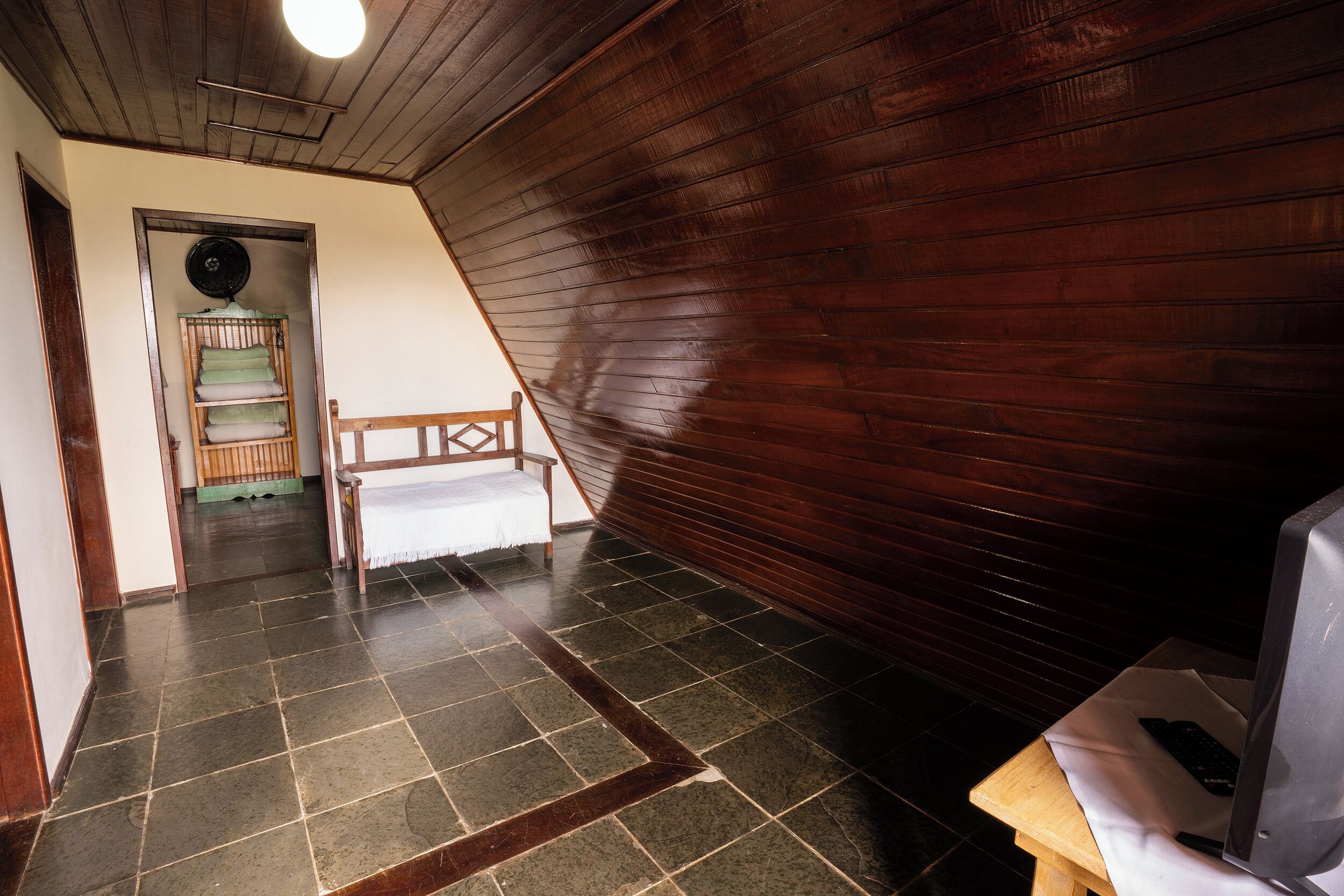 A room with a sloped ceiling, a wooden bench, a television on a table, and a fan on the wall.