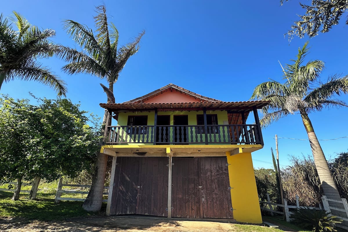 Uma casa de dois andares com varanda, cercada por palmeiras e um muro de madeira.