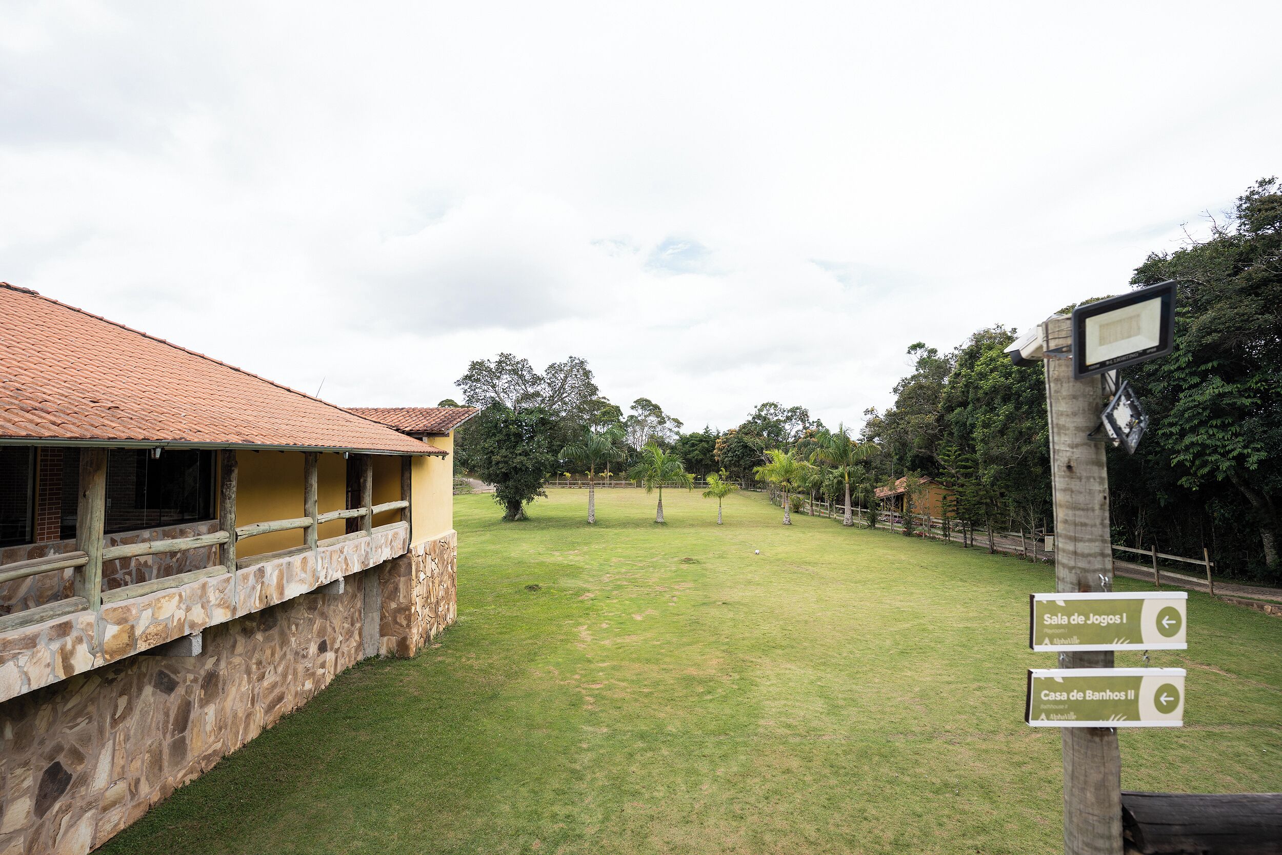 A courtyard with a stone wall, a yellow building, and directional signs for 'Sala de Jogos' and 'Casa de Banhos.'