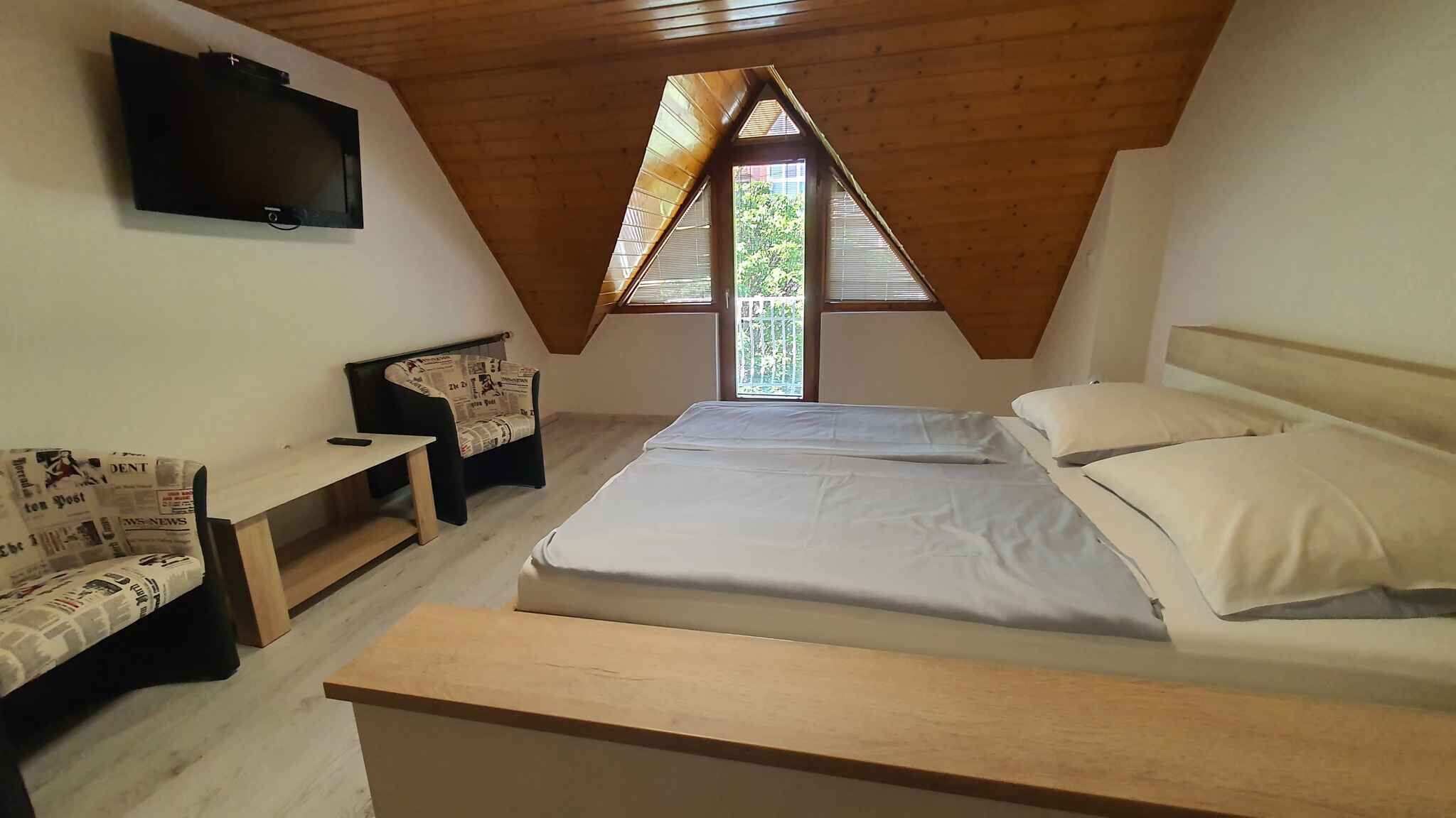 Une chambre avec un plafond en pente, un lit recouvert de draps blancs, une petite table, une chaise avec un coussin à motifs de journal et un téléviseur fixé au mur.