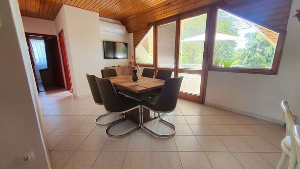 Un coin repas avec une table et des chaises en bois, un téléviseur fixé au mur et de grandes fenêtres donnant sur un paysage verdoyant.