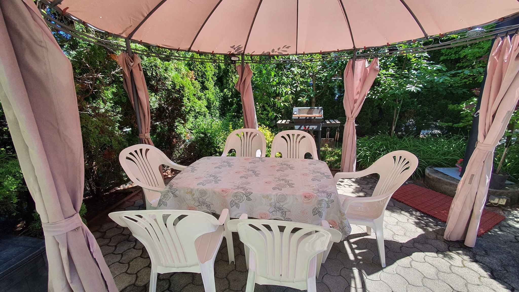 A covered outdoor seating area with a table and chairs, surrounded by greenery.