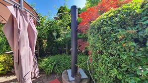 Un jardin avec un grand poteau noir, une pergola recouverte d’un tissu rose et diverses plantes vertes et rouges.