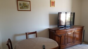 Een eethoek met een ronde tafel, bedekt met een beige tafelkleed, twee houten stoelen, een houten kast met een televisie erop, en twee ingelijste foto’s aan de muur.