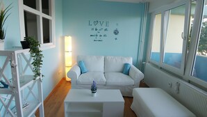 Una habitación con un sofá blanco, una mesa de centro blanca y un banco blanco. Se observa una pared con una frase decorativa y una ventana.