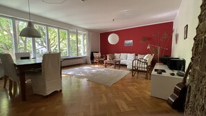 Uma sala de estar espaçosa com uma janela ampla, um sofá branco, uma mesa de centro de madeira e um espaço de jantar com cadeiras e mesa.