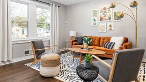 Ein modernes Wohnzimmer mit einer Couch, Sesseln, einem Couchtisch und einem großen Fenster mit Vorhängen.