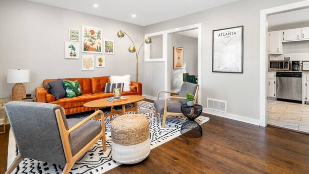 Ein modernes Wohnzimmer mit einer Couch, Sesseln, einem Couchtisch und einem Teppich. Im Hintergrund sind Wandbilder und eine Küche zu sehen.