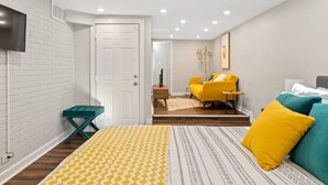 Ein Schlafzimmer mit einem Bett, einem gelben Sofa, einem türkisfarbenen Beistelltisch und einem an der Wand montierten Fernseher.
