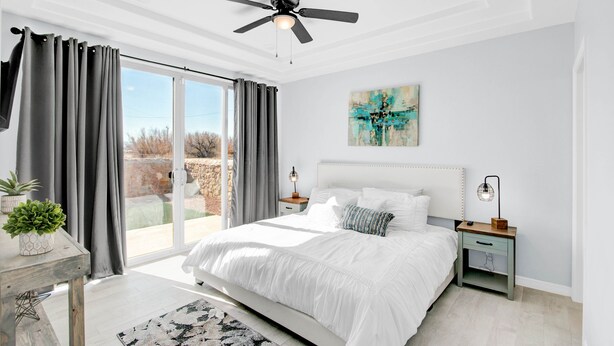 Une chambre avec un grand lit, un ventilateur de plafond, une table de chevet avec une lampe et une vue sur l’extérieur, accessible par une porte-fenêtre coulissante.