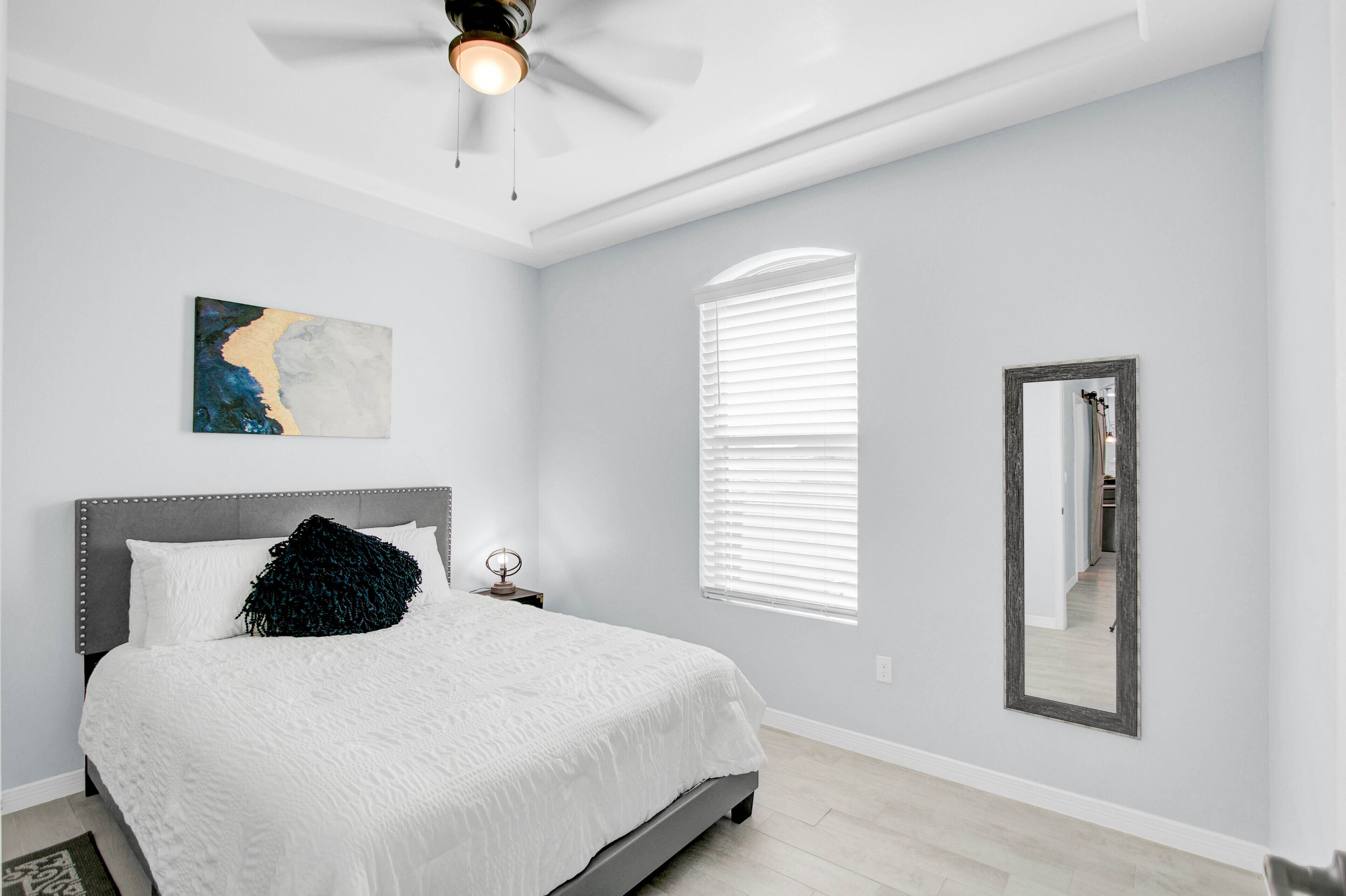 A bedroom with a bed, a ceiling fan, a window with blinds, a wall-mounted mirror, and a painting.