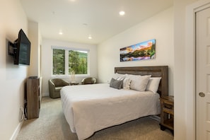 Une chambre avec un grand lit, un téléviseur fixé au mur, une fenêtre donnant sur un paysage verdoyant et un tableau au-dessus du lit.