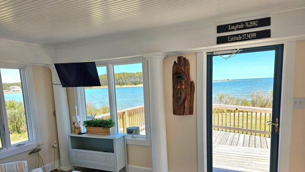 Une chambre avec vue sur l’eau, une terrasse en bois à l’extérieur, un téléviseur posé sur un meuble et un panneau indiquant la latitude et la longitude.