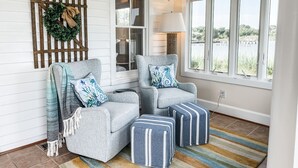 Une pièce chaleureuse avec deux fauteuils gris, un pouf rayé, une décoration murale en bois, une lampe et une vue sur un lac, visible à travers de grandes fenêtres.