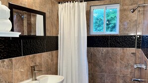 Une salle de bain équipée d’un lavabo, de toilettes, d’une douche et d’une fenêtre donnant sur un espace verdoyant.