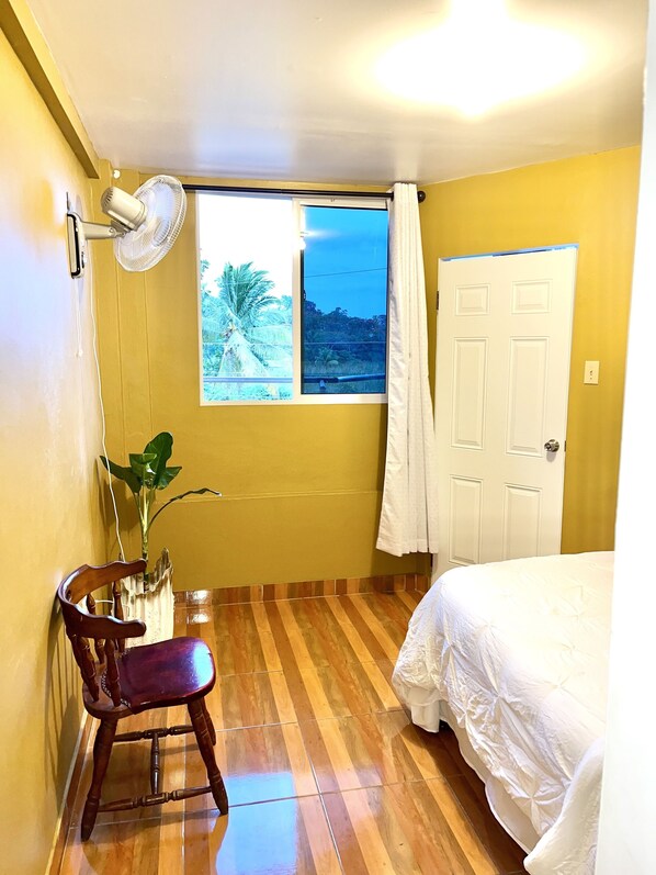 Quarto com cama, cadeira, janela vista para palmeiras, ventilador e porta.