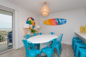 A dining area with a round table surrounded by blue chairs, a colorful surfboard art piece, and a beach view through a sliding glass door. - Beautiful Ocean views, Just renovated 2 bedroom 2 bath Condo, 2 parking spots (Ocean City)