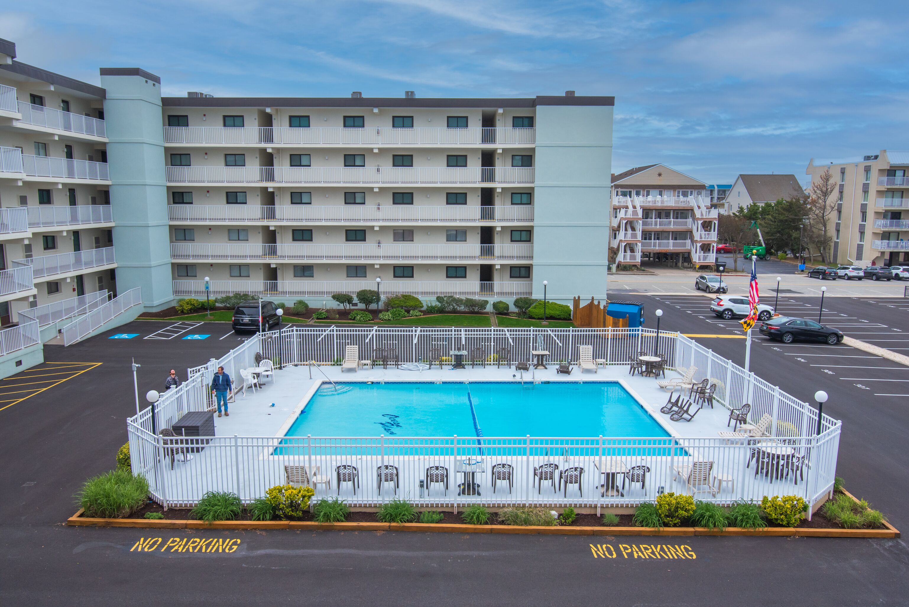 A hotel with a swimming pool surrounded by a white fence, outdoor seating, and a parking area marked "NO PARKING".